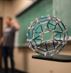 A lecturer lectures in the background and in the foreground stands a three-dimensional object.