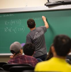 A lecturer writes on a chalkboard as students look on.