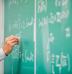 An instructor writes mathematics formulas on a green chalkboard.