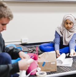 Students sit on the ground and flip through a book and handle objects during an activity.